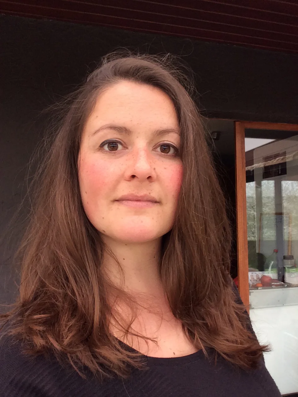 Portrait en gros plan d'une femme aux cheveux bruns longs, vêtue d'un haut noir, devant une structure architecturale en bois et verre.